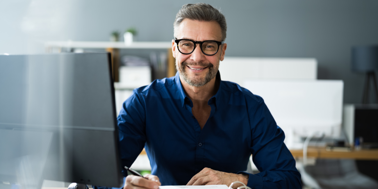 Smiling man in glasses sit at table smiling while looking at camera in office.